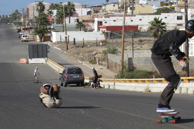 Eine Straße mit skatenden Menschen, geparkten Fahrzeugen und Häusern und Bäumen im Hintergrund.