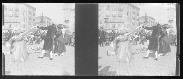 Schwarzes und weißes Foto eines Faschingsumzugs mit tanzenden Menschen in Kostümen auf der Straße, umgeben von Gebäuden, Straßenlaternen und Straßenlaternen unter einem klaren Himmel.