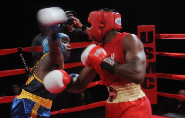 Zwei Boxer im Ring bei einem Kampf, ein Fotograf hält die Action im Hintergrund fest, mit einem kleinen Publikum im Dunkeln.