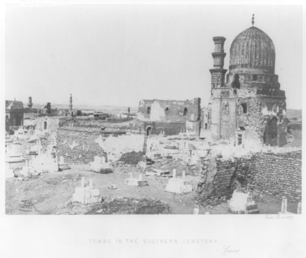 Schwarzes und weißes Foto eines Friedhofs mit Grabsteinen und einer Moschee im Hintergrund, mit Text unten.