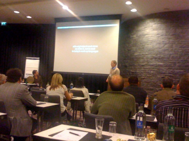 Ein Mann präsentiert mit einem Bildschirm hinter ihm, während eine Gruppe von Menschen zuhört.