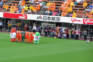 Ein Fußballspiel in einem Stadion mit sechs Spielern, drei Fußballen, vielen Zuschauern in Regenschutzjacken mit Schirmen und mehreren Kameraleuten.