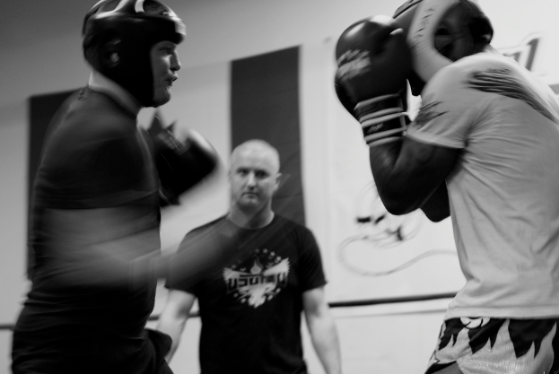 Zwei Boxer mit Helmen und Handschuhen kämpfen im Ring, mit einem Schiedsrichter zwischen ihnen und einem Banner an der Wand im Hintergrund.