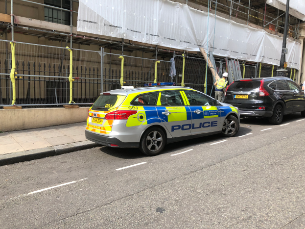 Ein Polizeiwagen steht an der Straße neben einem Gebäude, zwei Helme tragende Personen stehen daneben; im Vordergrund sind ein Geländer und ein Pfahl zu sehen, im Hintergrund ist ein weißes Tuch über das Gebäude drapiert, was auf eine polizeiliche Untersuchung nach einem Messerangriff in London hindeutet.