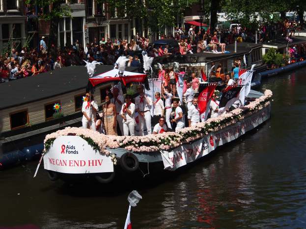 Eine Gruppe von Menschen auf einem Boot im Wasser, umgeben von Fahnen, Blumen und anderen Gegenständen, mit Gebäuden, Bäumen und einem Laternenpfahl im Hintergrund während des Amsterdam-Rot-Kreuz-Parades.
