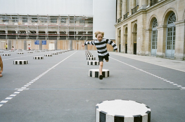 Ein Spielplatz mit vier Kindern, eines läuft auf schwarzen und weißen Betonstufen, umgeben von einem Schulgebäude.