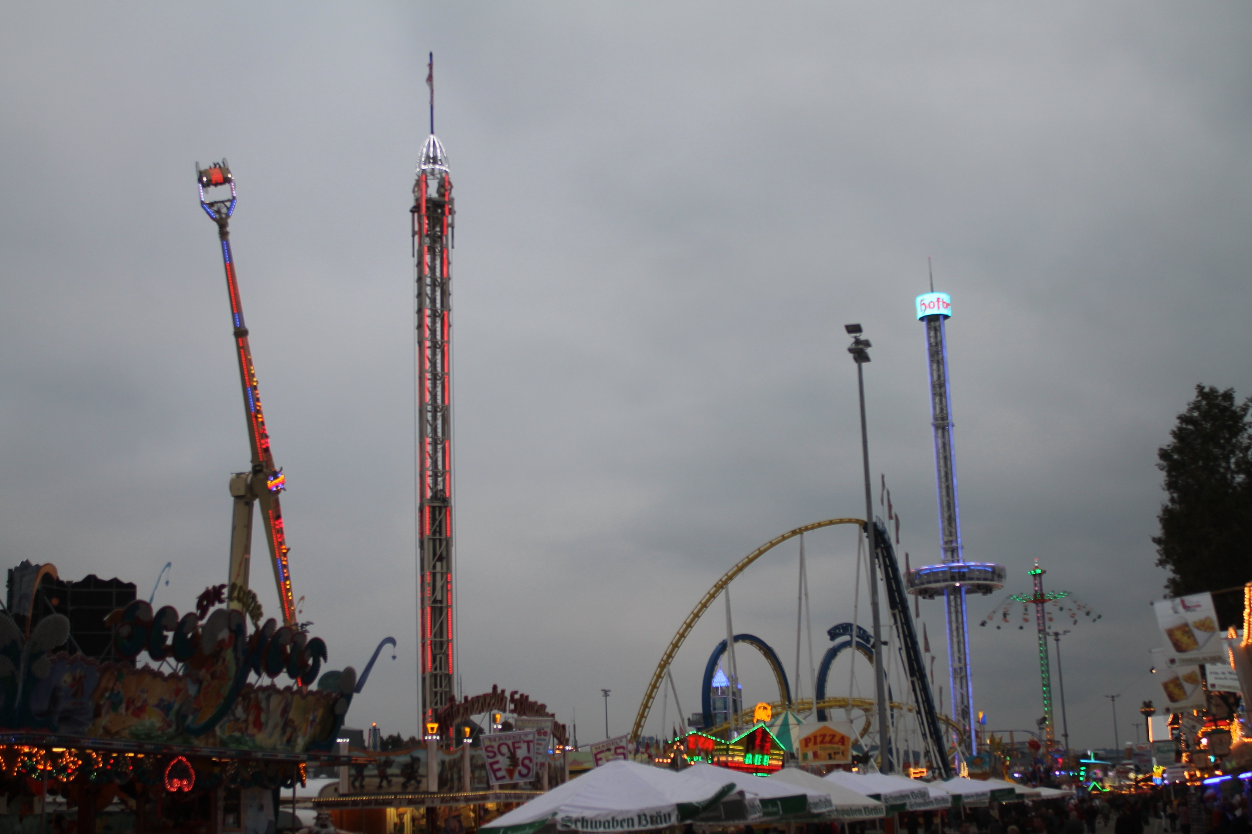 Eine nächtliche Faschingszene mit einer Menschenmenge, die sich um beleuchtete Fahrgeschäfte, Pfähle und Bäume versammelt hat, unter einem bewölkten Himmel.