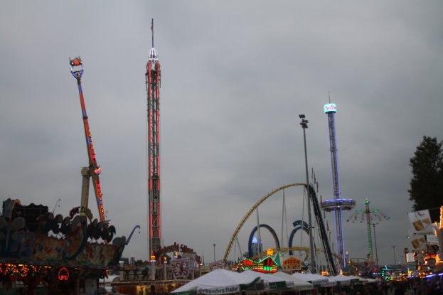 Eine nächtliche Faschingszene mit einer Menschenmenge, die sich um beleuchtete Fahrgeschäfte, Pfähle und Bäume versammelt hat, unter einem bewölkten Himmel.
