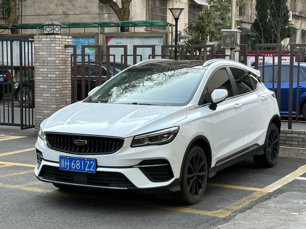 Ein weißer Volvo XC40 T5 AWD Inscription auf einem Parkplatz mit Gebäuden, Bäumen, Pfählen, Lampen und anderen Objekten im Hintergrund.