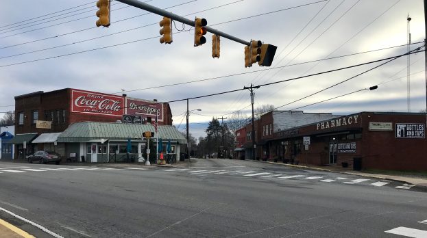 Eine Stadtstraßenkreuzung mit Ampel, Fahrzeugen, Gebäuden mit Namensschildern, Strommasten mit Drähten, Bäumen und einem bewölkten Himmel.