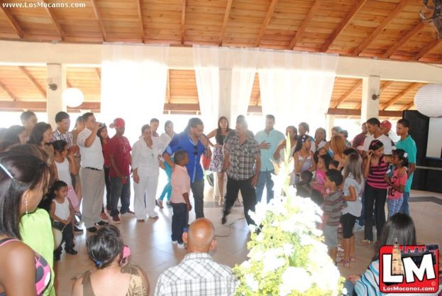 Eine Gruppe von Menschen mit einer Pflanze und einem Logo unten, Vorhängen und einem Lautsprecher im Hintergrund sowie Text in der oberen linken Ecke.